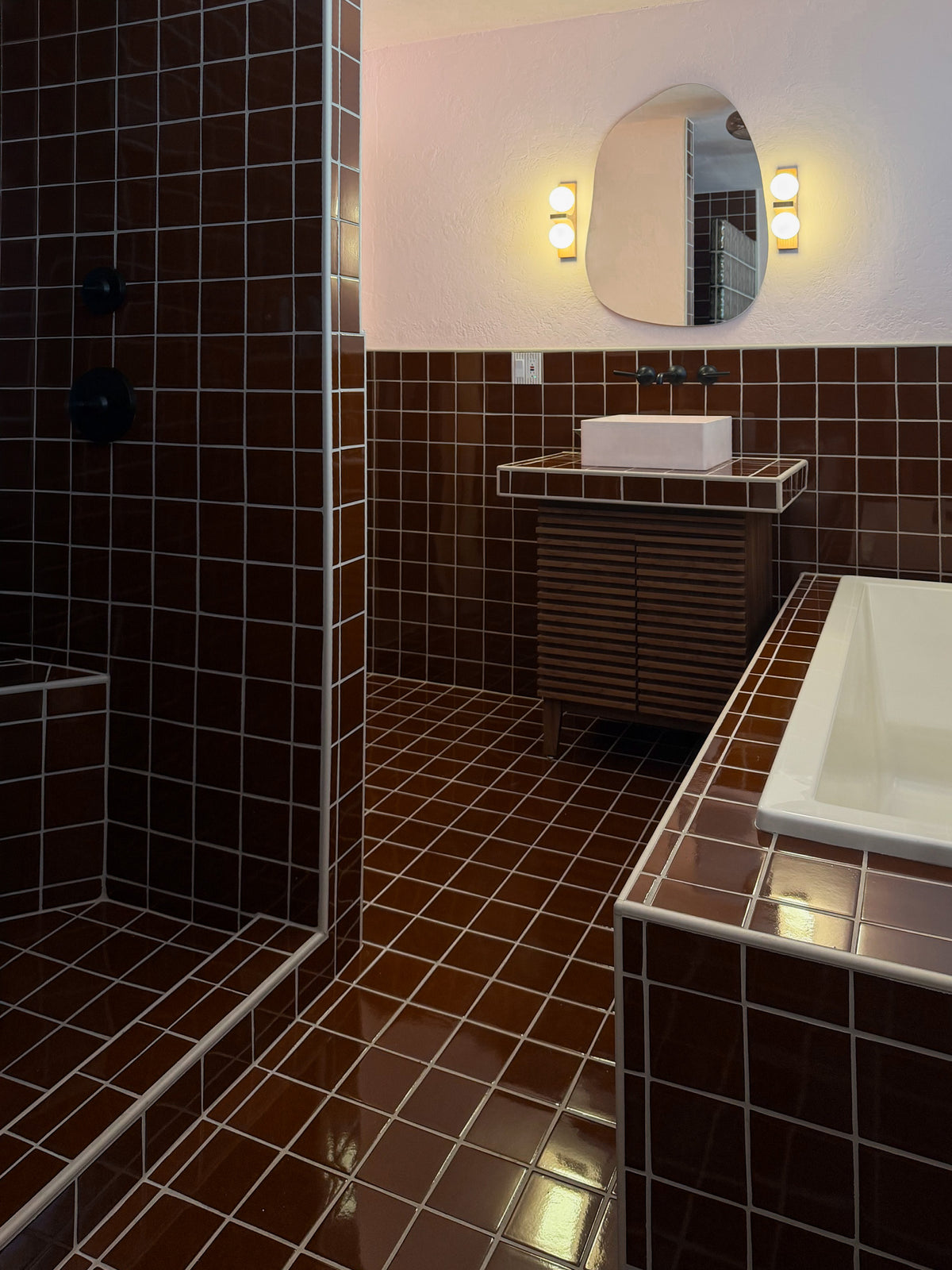 A modern bathroom interior with dark brown glossy square tiles, a white rectangular sink, a matching tiled vanity, an oval mirror with wall lights, and a shower area on the left.