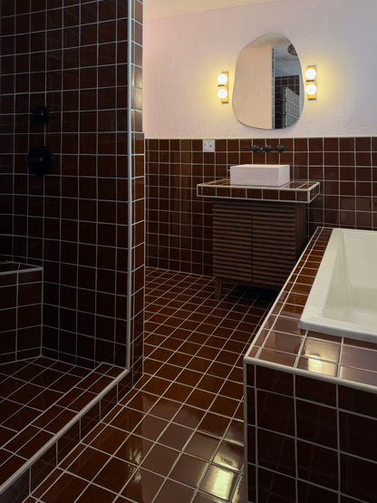 A modern bathroom interior with dark brown glossy square tiles, a white rectangular sink, a matching tiled vanity, an oval mirror with wall lights, and a shower area on the left.
