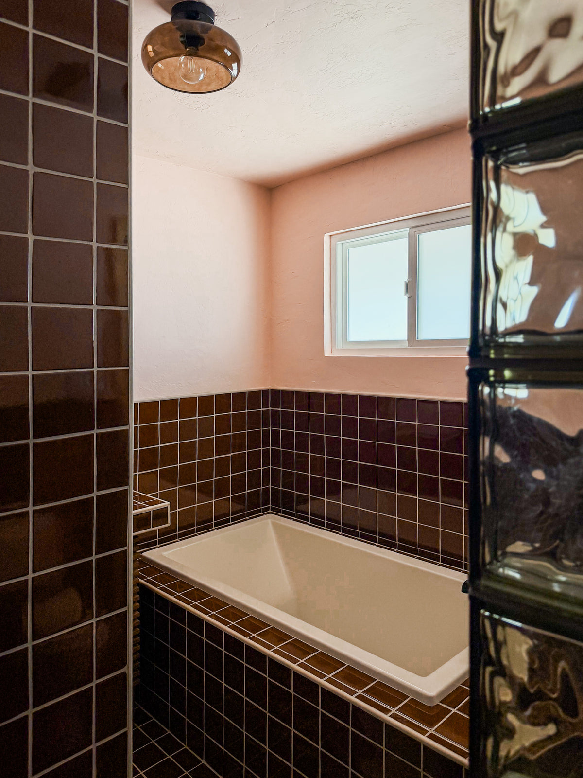 A bathroom interior with a built-in white bathtub surrounded by dark brown glossy tile, a peach-pink wall, a small window, and a hanging amber glass pendant light.