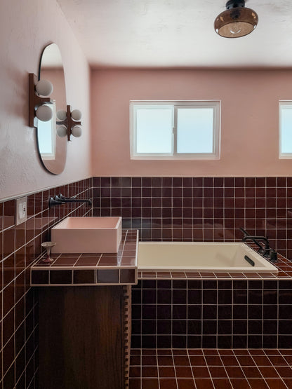 A retro-style bathroom interior featuring dark brown glossy tile walls and floor, a pink vessel sink on a matching tiled vanity, a cream bathtub, a round wall mirror, and a window.