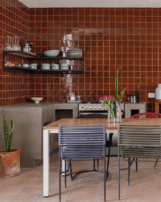 A rustic kitchen corner with glossy terracotta brown wall tiles, concrete countertops, a built-in stove, and a wooden dining table surrounded by modern slatted chairs in blue, green, and red.