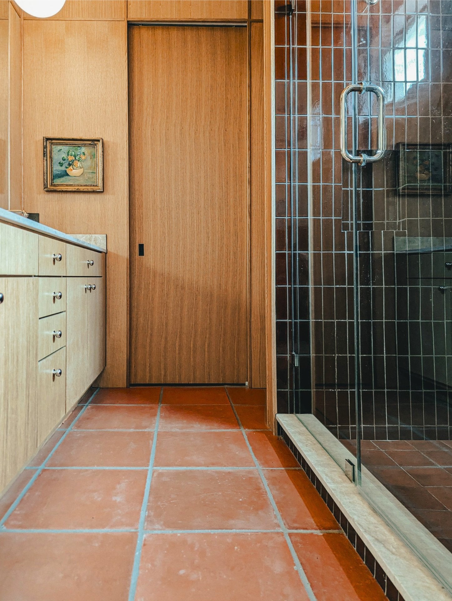 A modern bathroom interior with terracotta tile flooring, light wood cabinets and door, a glass shower enclosure with a metal handle, and a small framed painting on the wall.
