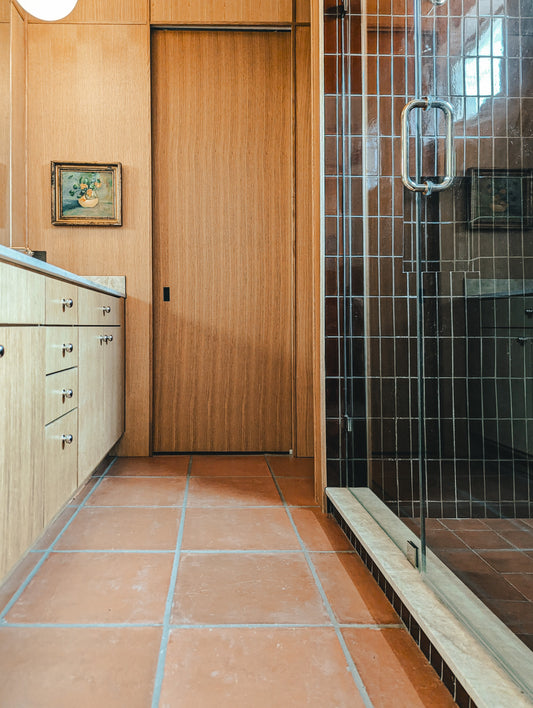 A modern bathroom interior with terracotta tile flooring, light wood cabinets and door, a glass shower enclosure with a metal handle, and a small framed painting on the wall.