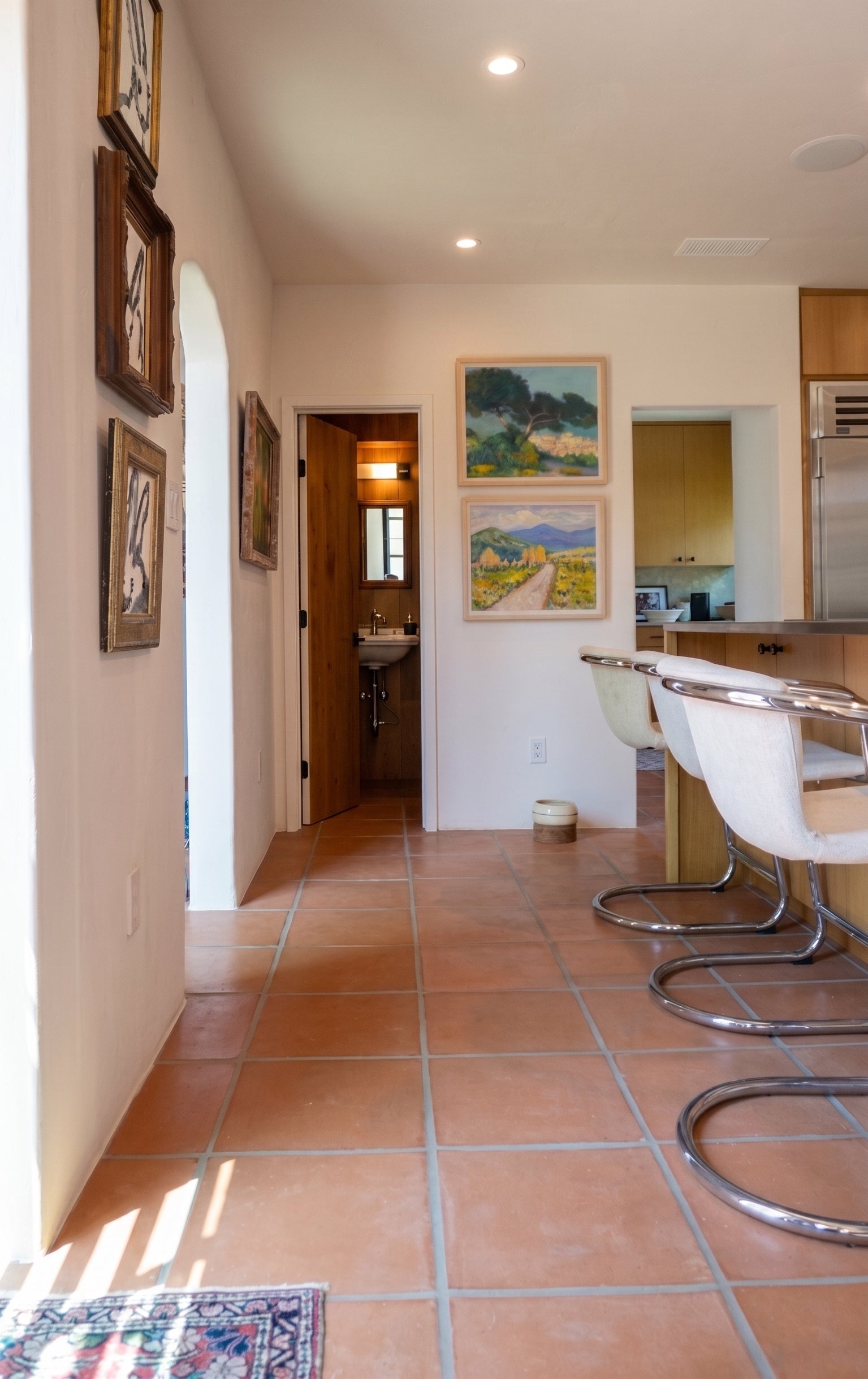 A hallway in a home with terracotta tile floors, cream walls, framed artwork, and a kitchen area with white bar stools on the right.