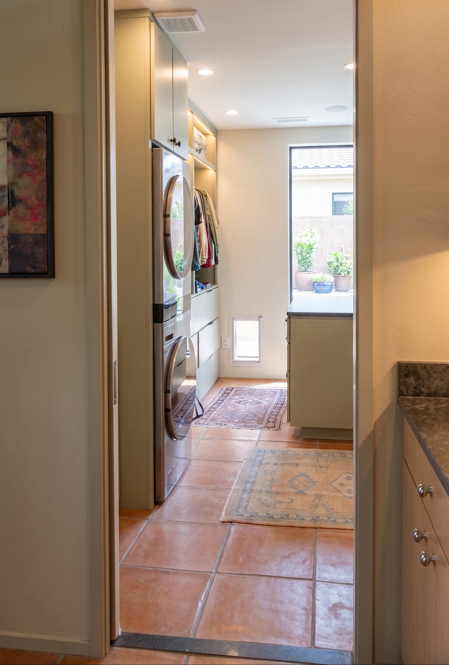 An open doorway looking into a compact laundry room with a stacked washer and dryer, terracotta tile flooring, and a small window near the back.