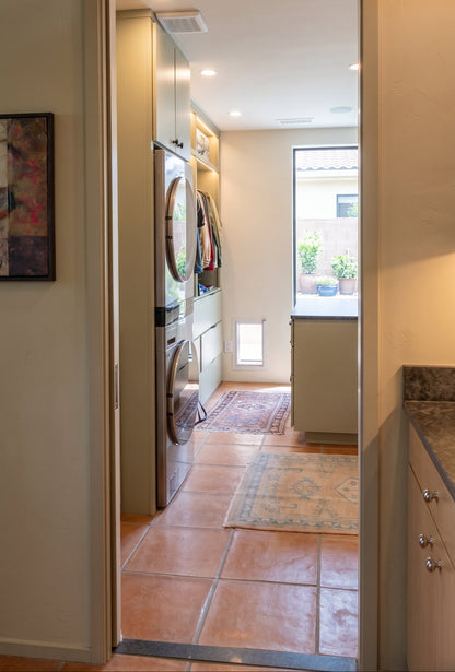 An open doorway looking into a compact laundry room with a stacked washer and dryer, terracotta tile flooring, and a small window near the back.