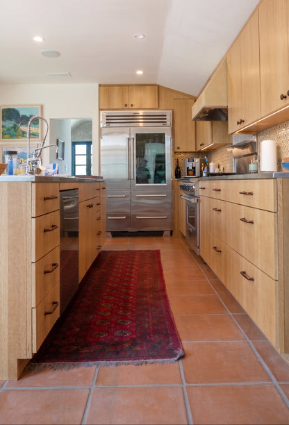 A long kitchen island with light wood cabinets, stainless steel appliances, terracotta tile flooring, and a red patterned runner rug.