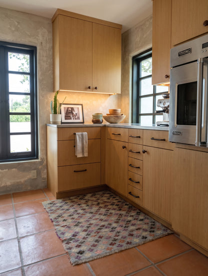 A kitchen interior with light wood cabinets, terracotta tile flooring, a patterned area rug, and built-in stainless steel appliances including a built-in oven. No person or animal is present.