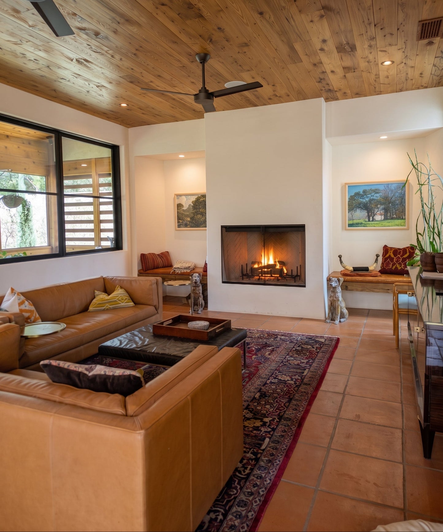 A warm, rustic living room with a tan leather sectional sofa, a brown leather loveseat, a patterned area rug, terracotta tile floors, and a built-in fireplace against a white wall.