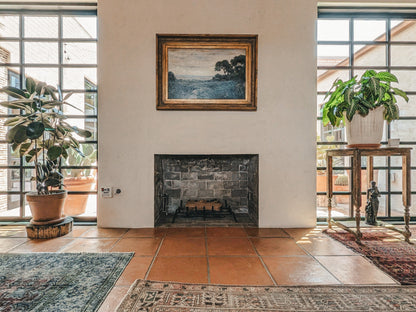 A framed landscape painting hangs on a white wall above a stone fireplace in a sunlit room with terracotta tile floors, large windows, potted plants, and patterned rugs.