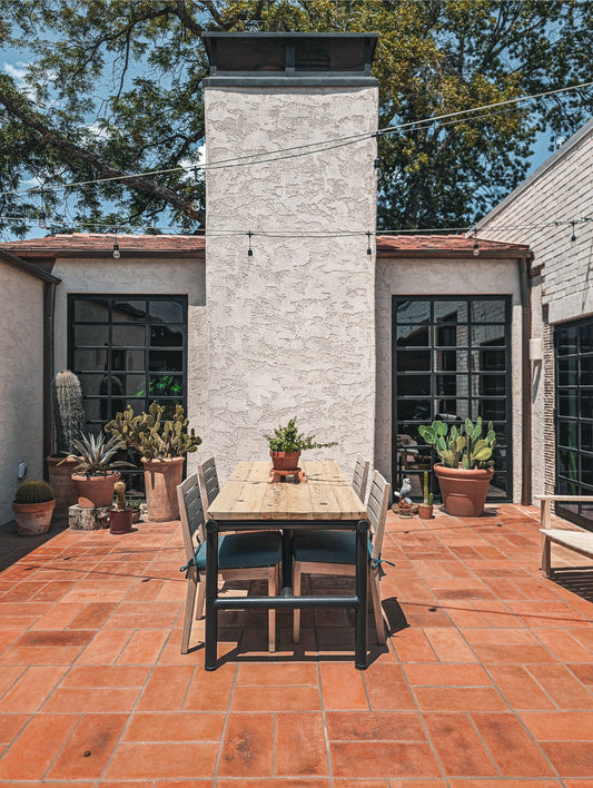 Outdoor patio dining setup with a rectangular wooden table and four chairs with blue cushions, set on terracotta tile in front of a stucco house with large black-framed windows and potted cacti.