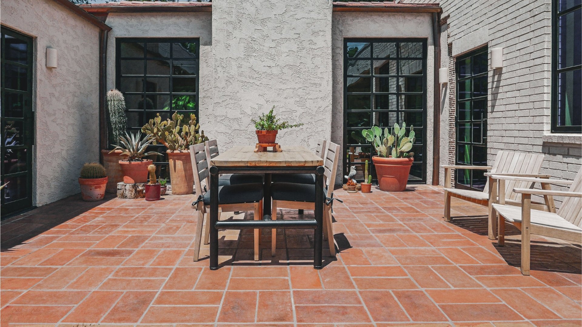 A modern outdoor patio dining set featuring a rectangular wooden table with a black metal frame and four black cushioned chairs, set on terracotta tile flooring next to white and gray stucco walls with potted plants.