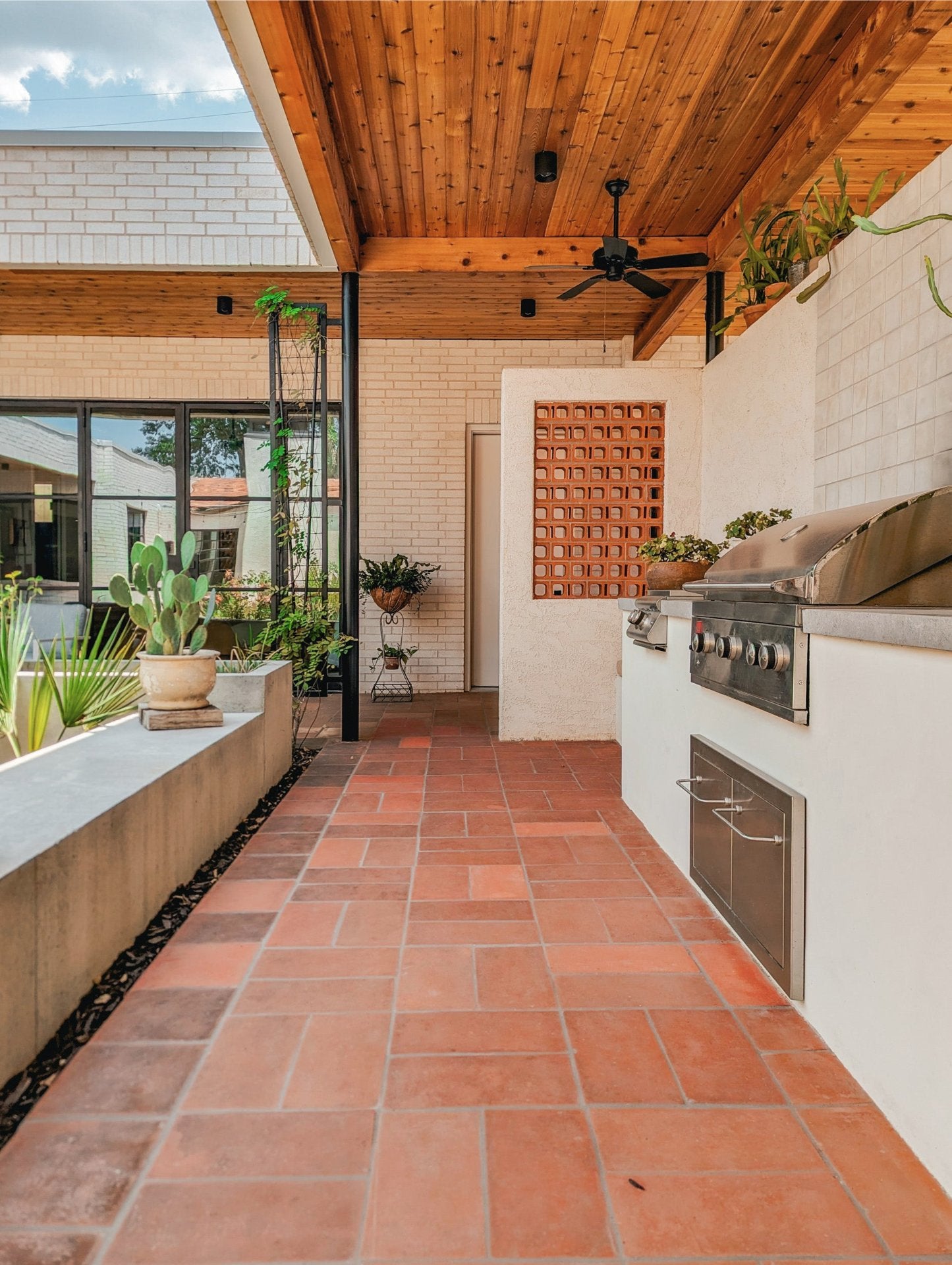 An outdoor kitchen area with a built-in stainless steel grill and cooktop on white cabinetry, terracotta tile flooring, a wooden ceiling, and potted plants along a raised planter ledge.
