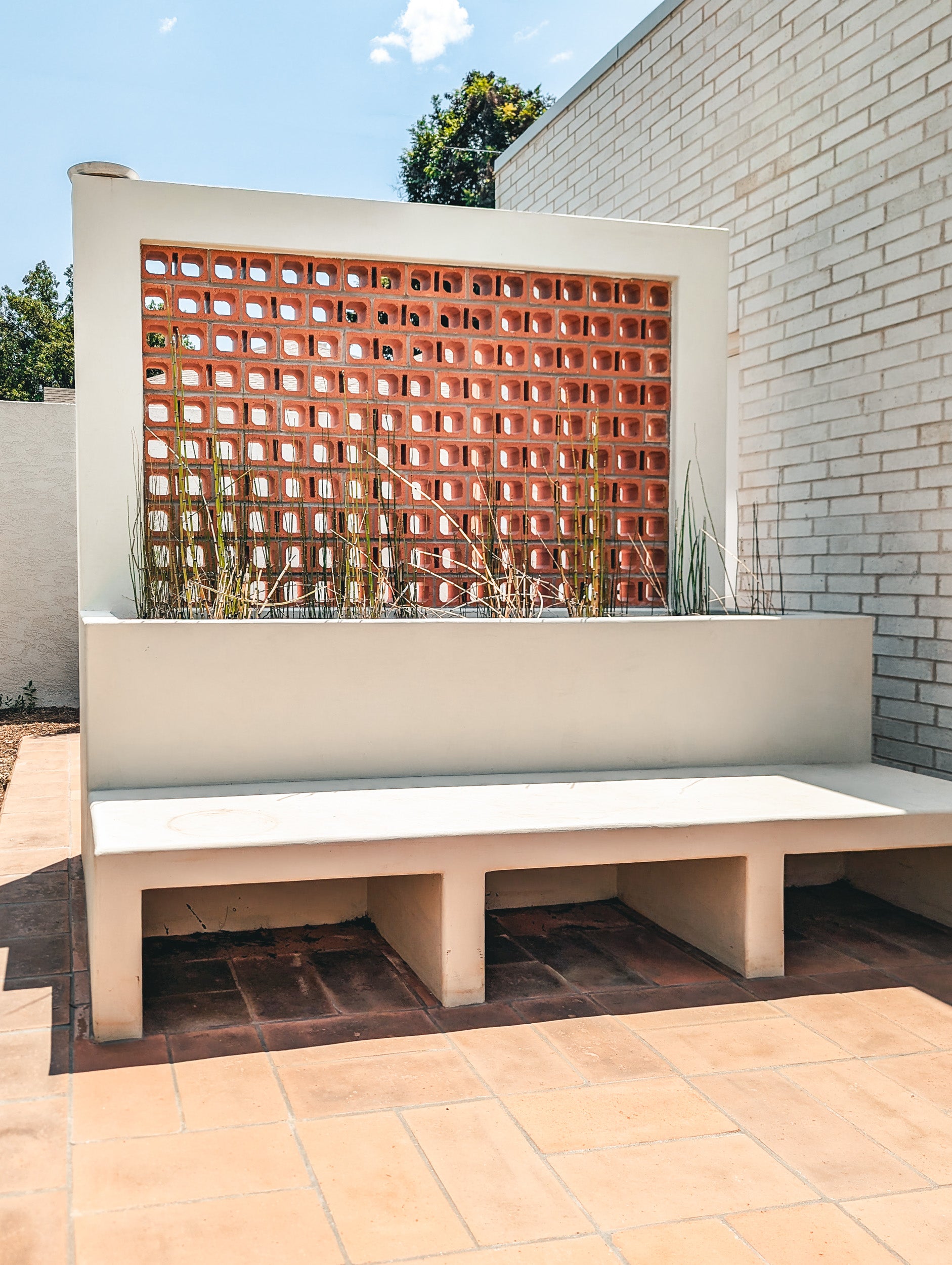 A modern outdoor concrete bench with a built-in planter box and terracotta brick block wall behind it, set on a tiled patio against a white brick building.