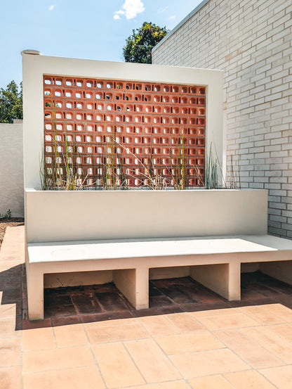 A modern outdoor concrete bench with a built-in planter box and terracotta brick block wall behind it, set on a tiled patio against a white brick building.