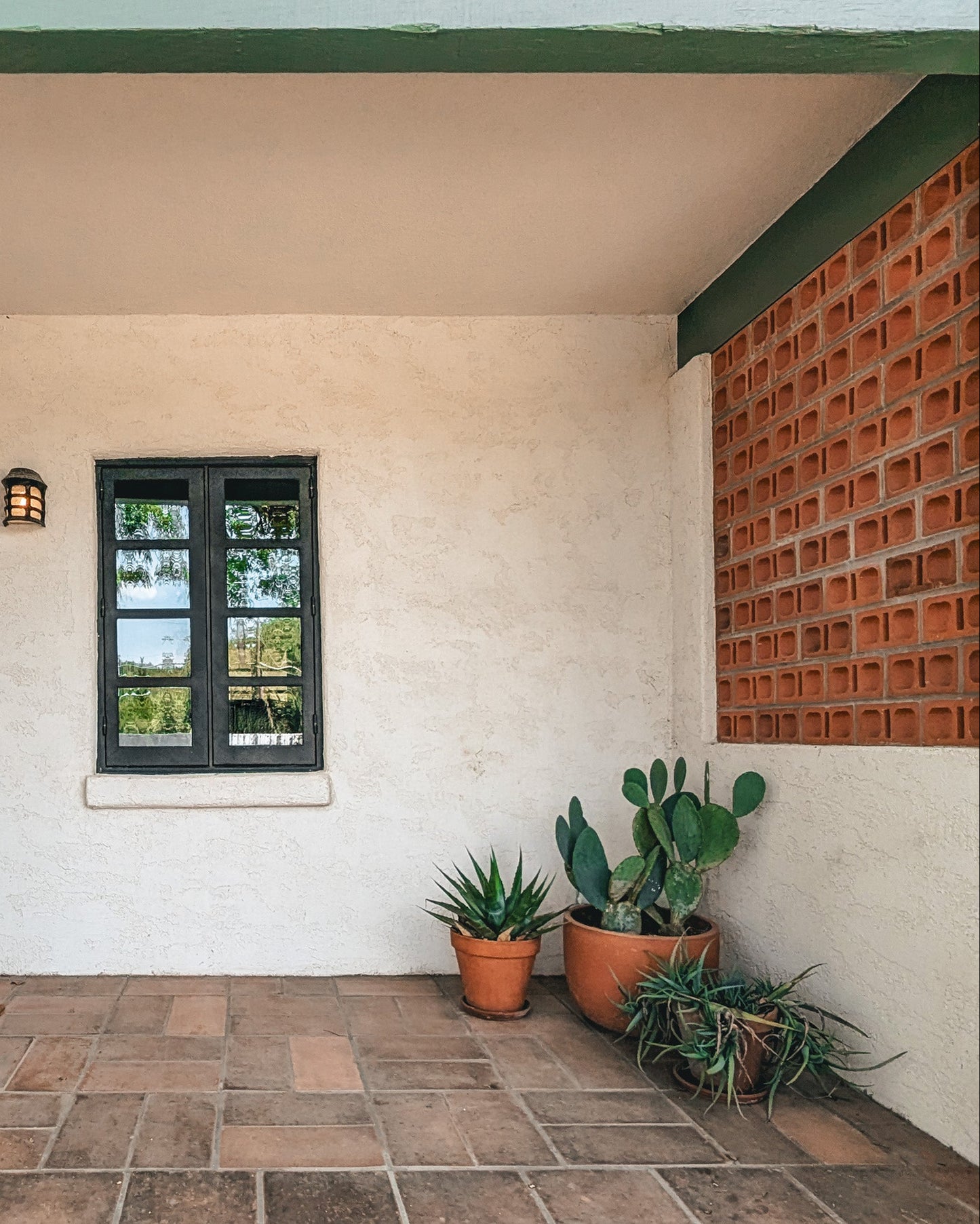 A front porch entryway with a stucco wall, tiled step, green door, black-framed window, terracotta brick wall, and potted plants.