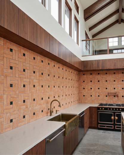 A modern kitchen interior with terracotta-colored geometric mosaic wall tiles, white countertops, a copper sink and faucet, dark wood cabinetry, and a black freestanding range with gold handles. High windows and exposed wood beams are visible above.