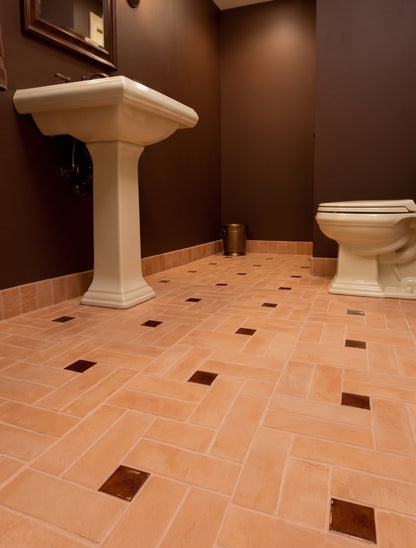 A bathroom interior with terracotta-colored herringbone floor tiles accented with small dark brown square inlays, dark brown walls, a white pedestal sink, and part of a white toilet.