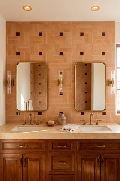 A double bathroom vanity with two sinks, rectangular mirrors, and a terracotta tile backsplash with small dark square accents. The countertop is a light beige stone, and the lower cabinets are dark wood with brass hardware.