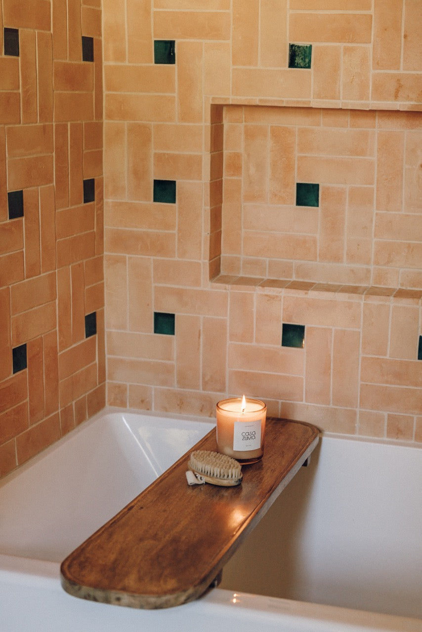 A lit candle in a glass jar sits on a wooden bath caddy draped over a white bathtub, with a small scrubber beside it. The background features warm terracotta tiled walls with small dark green square accents.