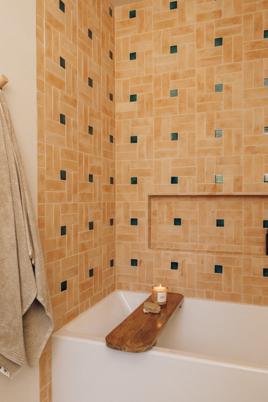 A bathroom corner with peach and beige mosaic tile walls, a white bathtub, a wooden tray on the tub edge, and a hanging towel.