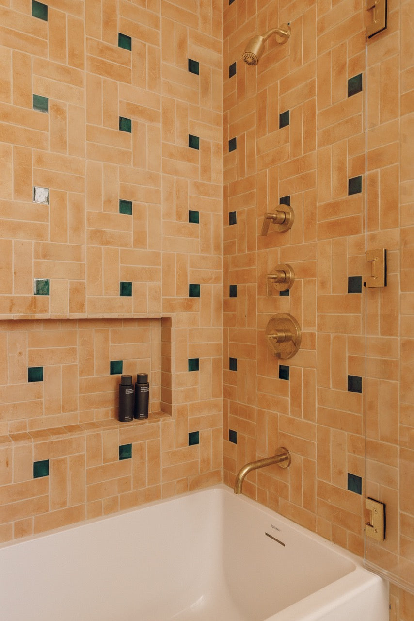 A close-up of a tiled bathroom corner with a white bathtub, warm beige and tan rectangular wall tiles, small dark green square accents, and gold-toned shower hardware.