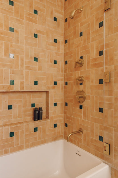 A close-up of a tiled bathroom corner with a white bathtub, warm beige and tan rectangular wall tiles, small dark green square accents, and gold-toned shower hardware.