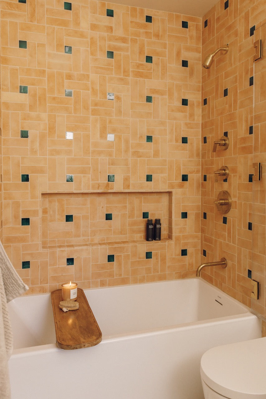 A bathroom interior with a white bathtub and shower area covered in warm beige mosaic tile walls with small dark green square accents. A wooden tray with a candle rests on the tub edge, and a toilet is partially visible in the lower right.