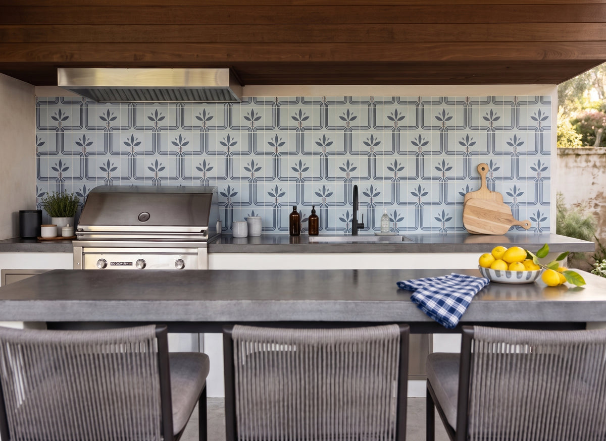 Outdoor kitchen scene with a patterned blue and white tile backsplash, stainless steel gas grill, dark countertop, bar stools, and a bowl of lemons on the counter.