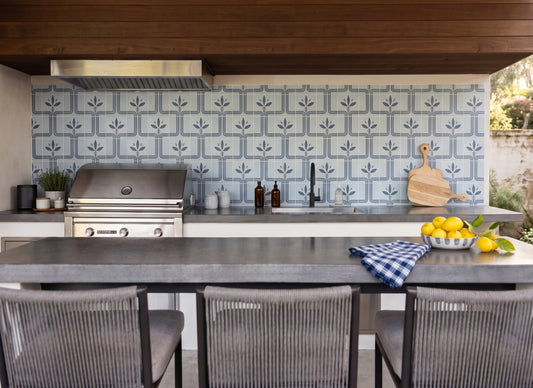Outdoor kitchen scene with a patterned blue and white tile backsplash, stainless steel gas grill, dark countertop, bar stools, and a bowl of lemons on the counter.
