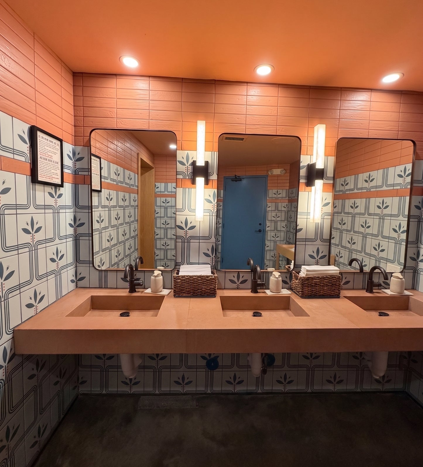 A modern three-sink bathroom vanity with a long wooden countertop, rectangular mirrors, patterned blue and white tiles, and warm orange walls and ceiling.