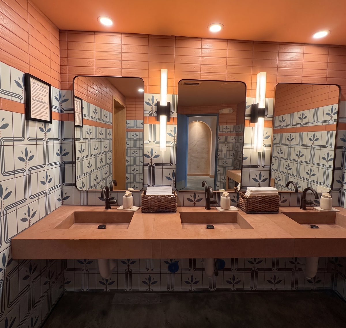 A modern commercial restroom features three peach-colored stone sinks set against walls decorated with coral subway tiles and blue-patterned retro floral tiles.
