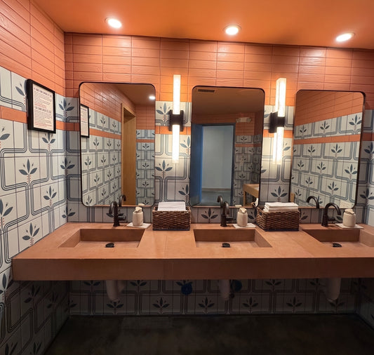 A modern double-sink restroom vanity with a light wood countertop, rectangular mirrors, patterned blue and white tiles, and warm orange walls and ceiling.