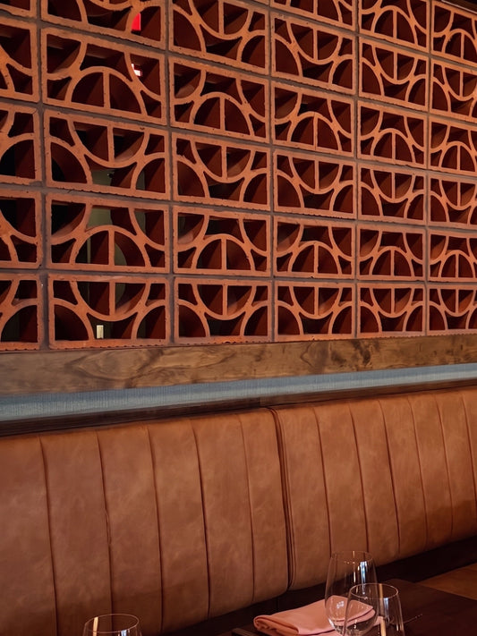 A warmly lit restaurant booth with a brown leather upholstered backrest, a decorative geometric wood screen behind it, and a dark wooden table set with pink folded napkins, clear wine glasses, and a small lit candle.