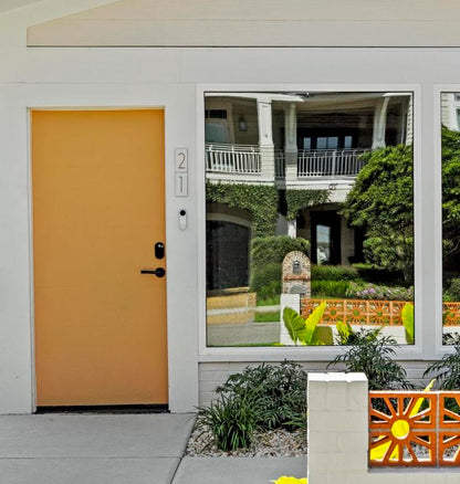 A modern house facade with a solid mustard-yellow front door on the left and a large white-framed window on the right reflecting a garden and building exterior.