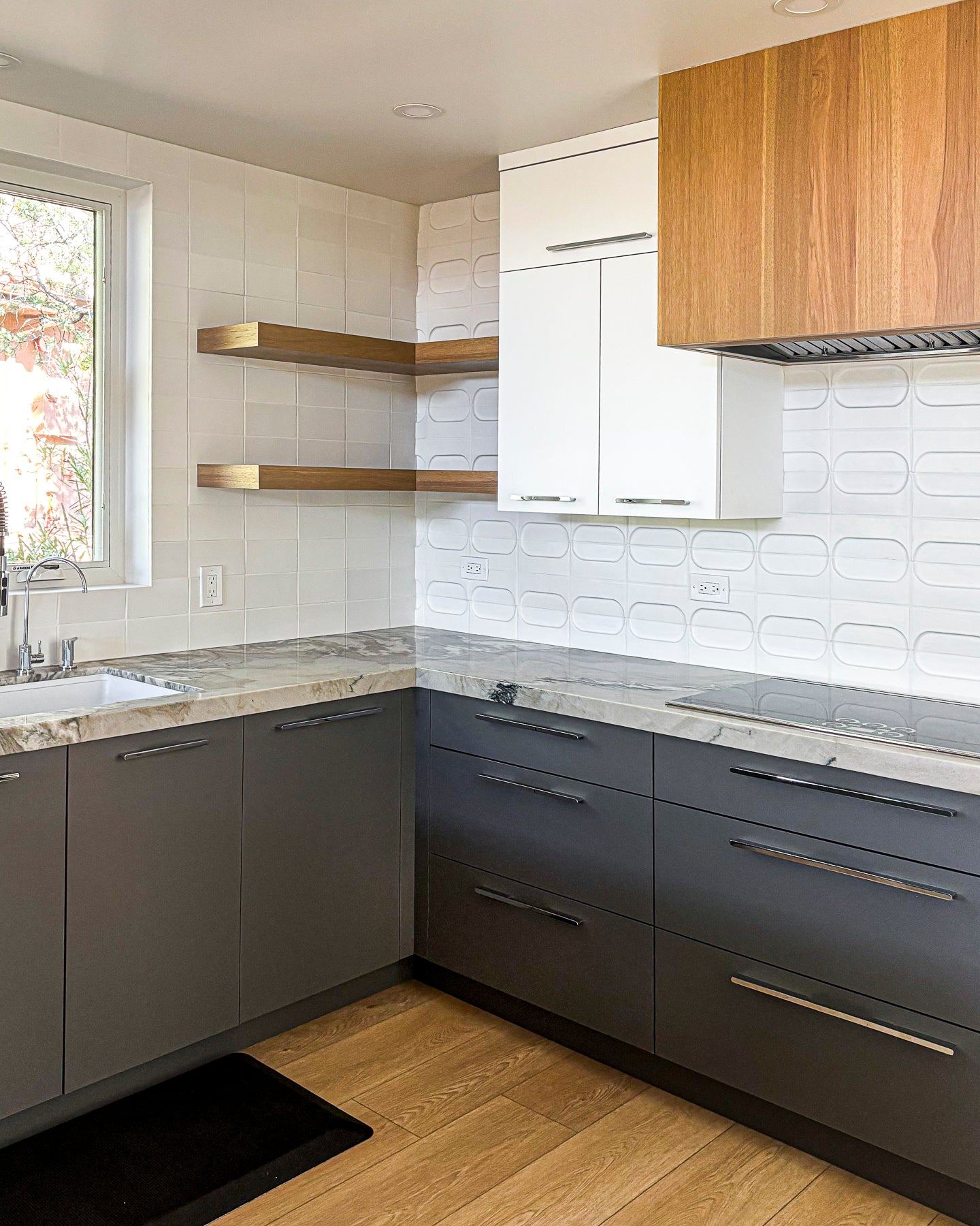 Modern kitchen cabinetry featuring dark gray lower cabinets, white upper cabinets, light wood floating shelves, a wood-look range hood, and marble countertops against a white textured tile backsplash.