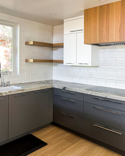 Modern kitchen cabinetry featuring dark gray lower cabinets, white upper cabinets, light wood floating shelves, a wood-look range hood, and marble countertops against a white textured tile backsplash.