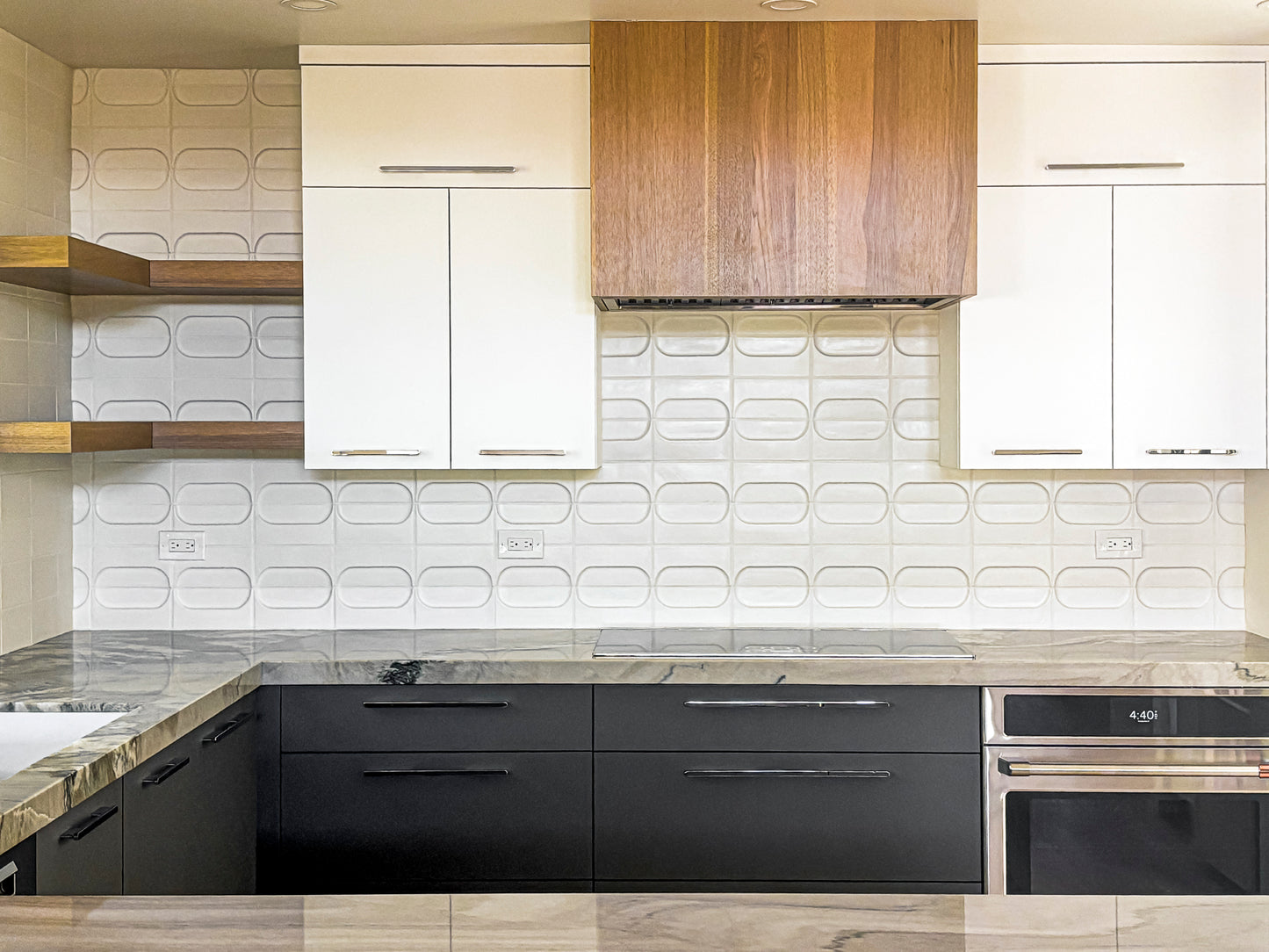 Modern kitchen interior with white upper cabinets, a wood-look range hood, black lower cabinets, a marble countertop, and a white textured tile backsplash.