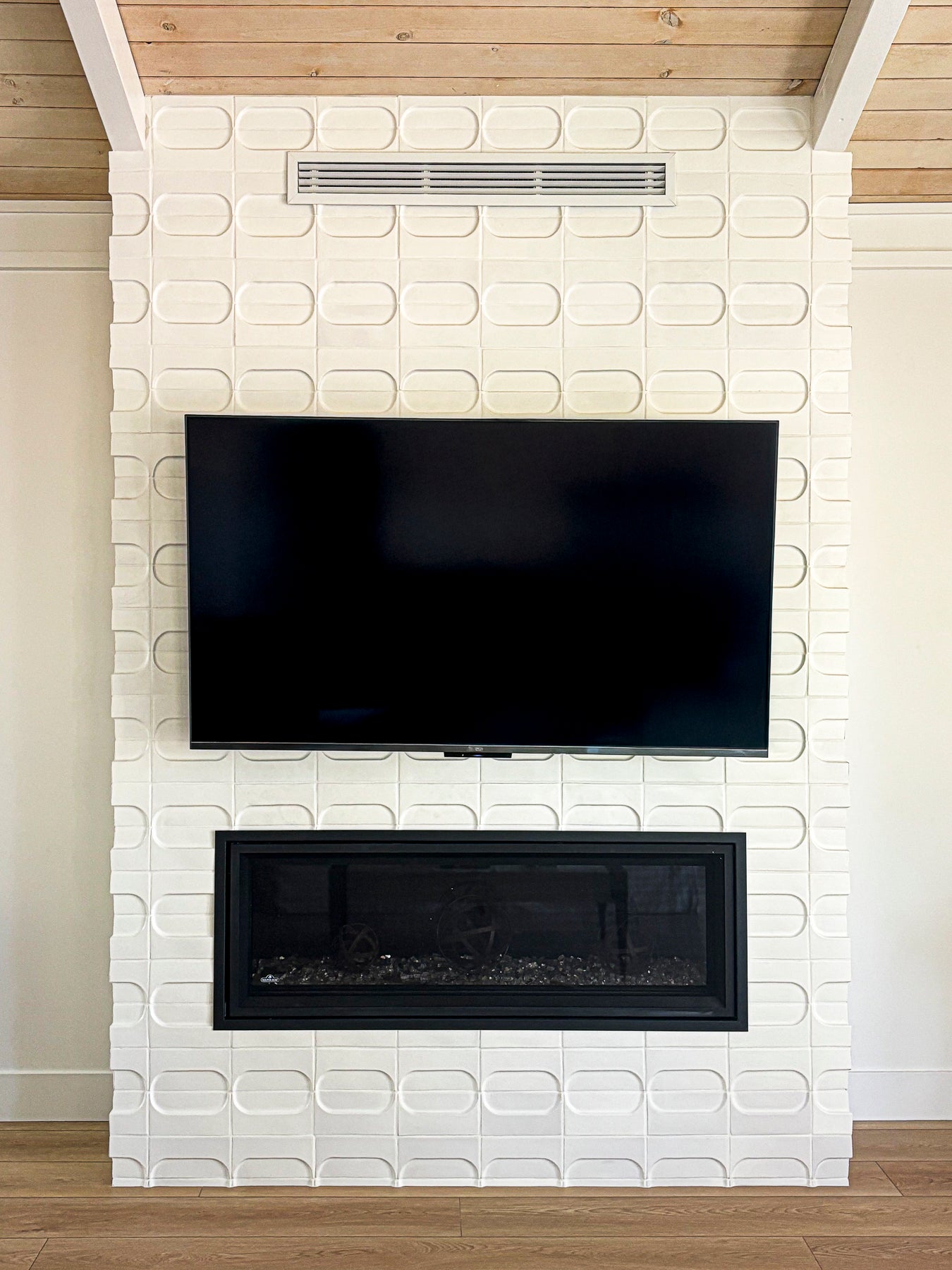 A modern white textured brick fireplace wall with a black flat-screen TV mounted above a black fireplace insert, set against a light wood ceiling and floor.
