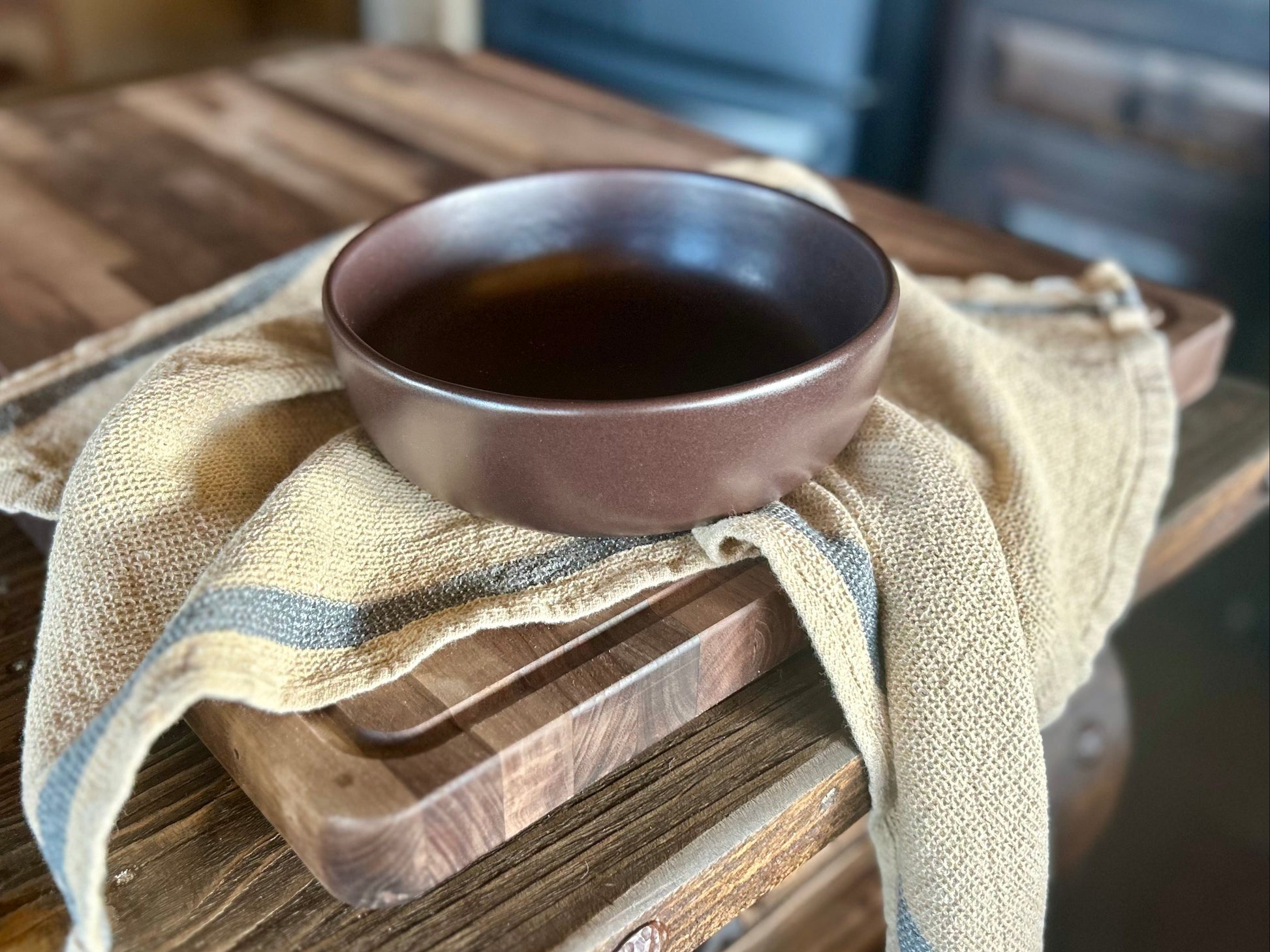 A small dark brown ceramic bowl filled with a dark liquid, resting on a rustic wooden cutting board draped with a beige linen cloth, set on a wooden table.