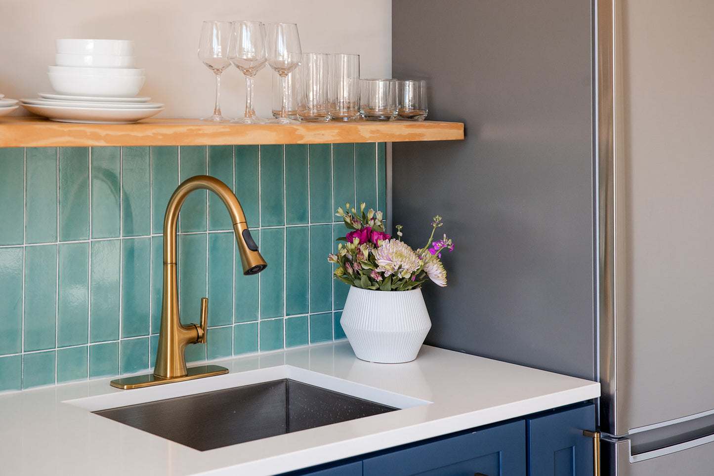 A modern kitchen corner featuring a matte brass gooseneck faucet, a white countertop with a dark inset sink, a teal tile backsplash, a wooden floating shelf with glassware, and a gray refrigerator.