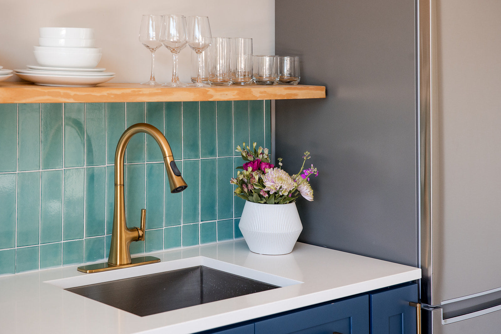 A modern kitchen corner featuring a matte brass gooseneck faucet, a white countertop with a dark inset sink, a teal tile backsplash, a wooden floating shelf with glassware, and a gray refrigerator.