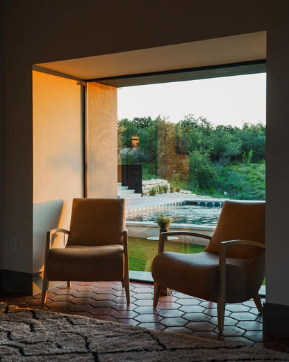 Two tan armchairs sit in a modern living room with a large picture window framing a view of a backyard swimming pool and lush green trees at sunset.