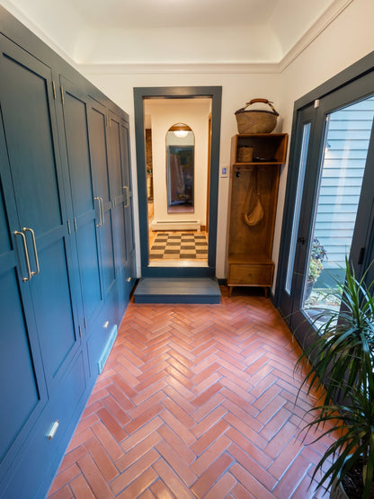 A bright entryway features terracotta herringbone tile floors, floor-to-ceiling dark blue storage cabinets, and a wooden coat rack next to a glass exterior door.