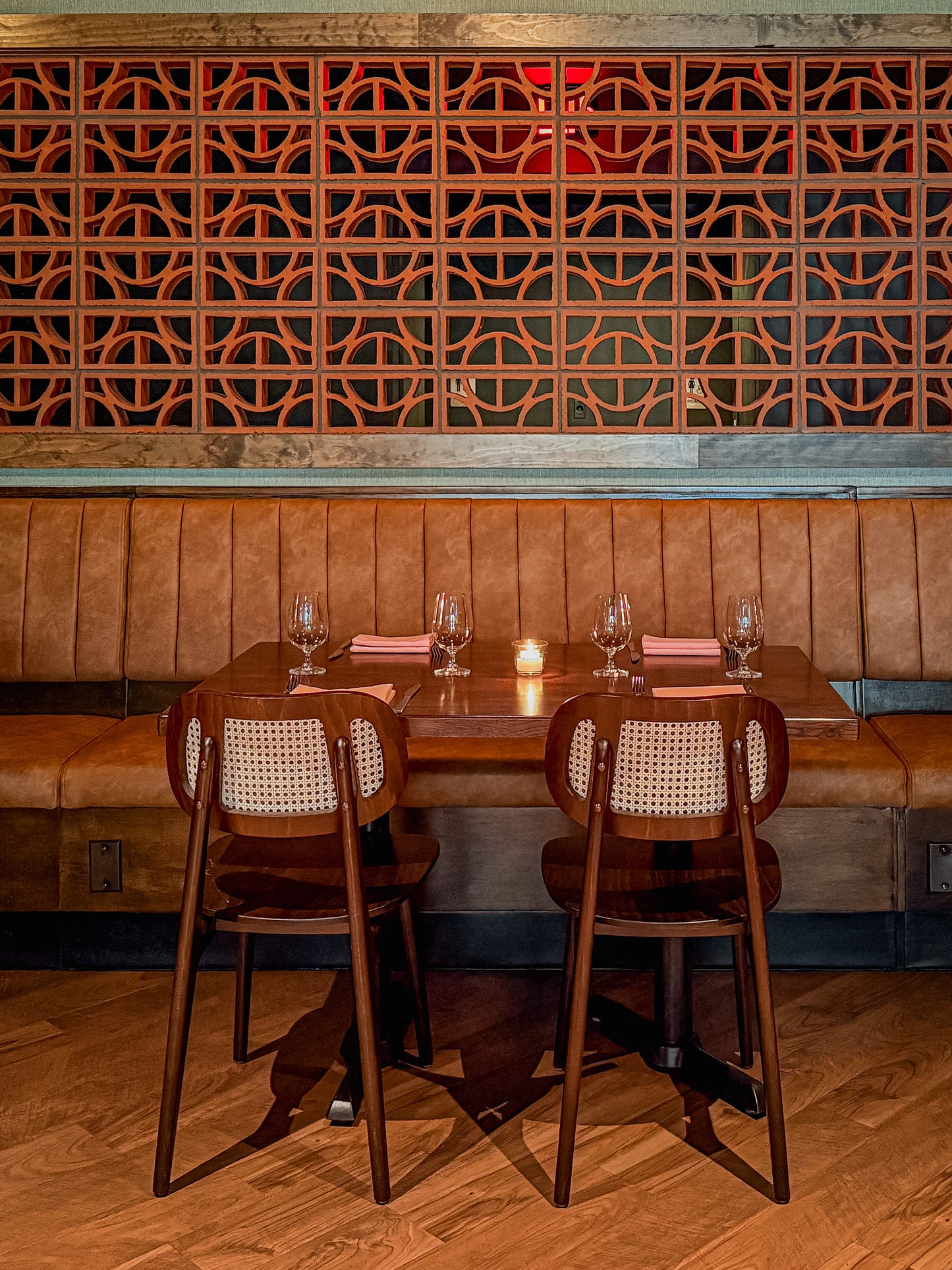 A restaurant booth setup with two dark wood chairs featuring woven cane backrests, set around a wooden table with wine glasses and pink napkins, against a brown leather booth and a decorative geometric wooden wall.