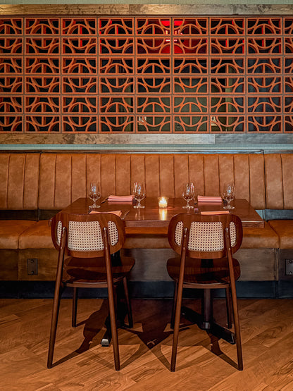 A restaurant booth setup with two dark wood chairs featuring woven cane backrests, set around a wooden table with wine glasses and pink napkins, against a brown leather booth and a decorative geometric wooden wall.