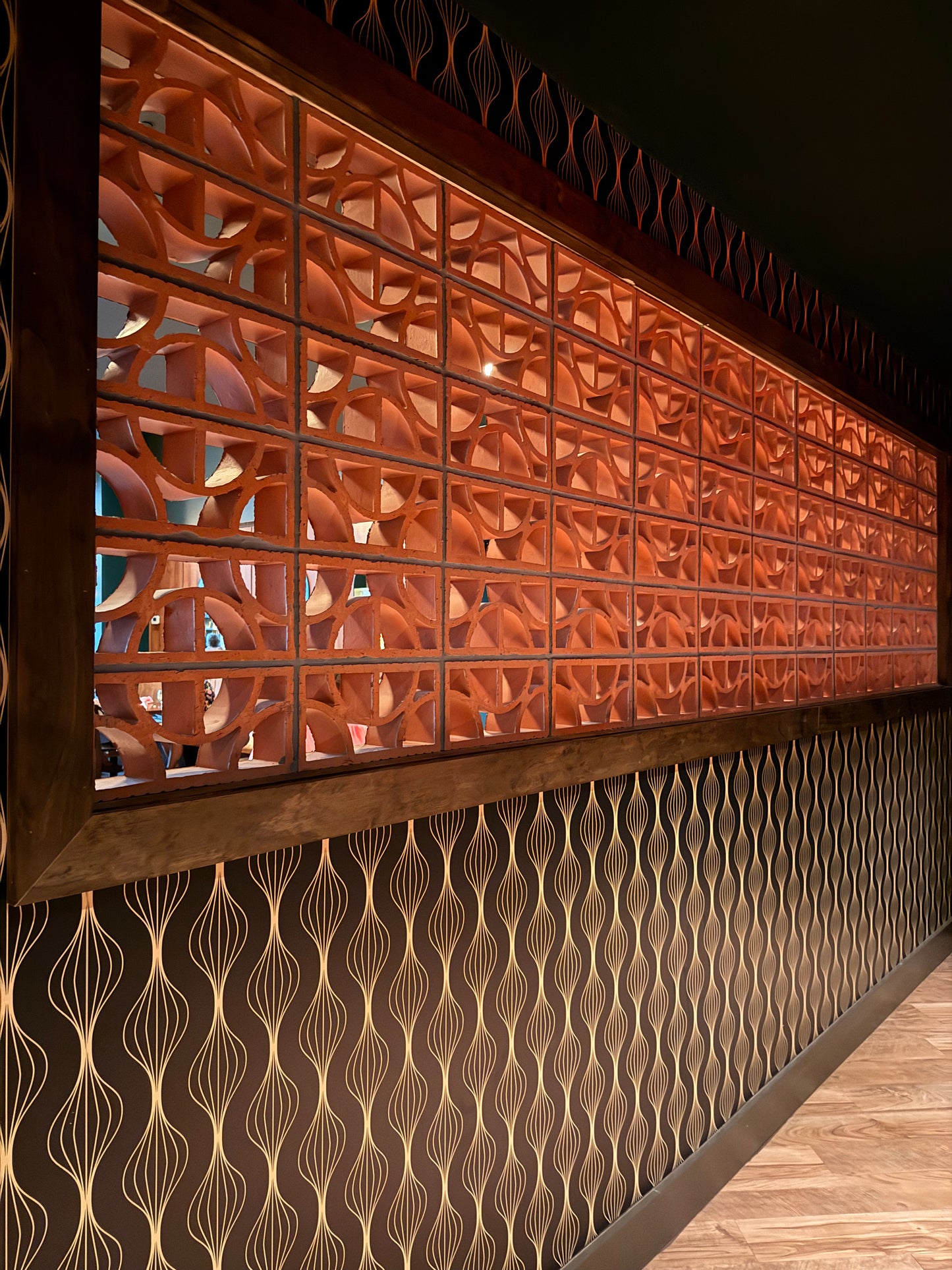 A long decorative wall panel with a warm copper-orange geometric screen pattern above a dark brown wavy patterned lower section, shown in an interior setting with wood flooring.