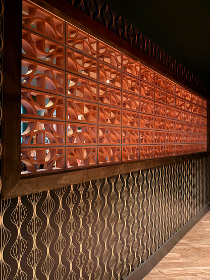 A long decorative wall panel with a warm copper-orange geometric screen pattern above a dark brown wavy patterned lower section, shown in an interior setting with wood flooring.