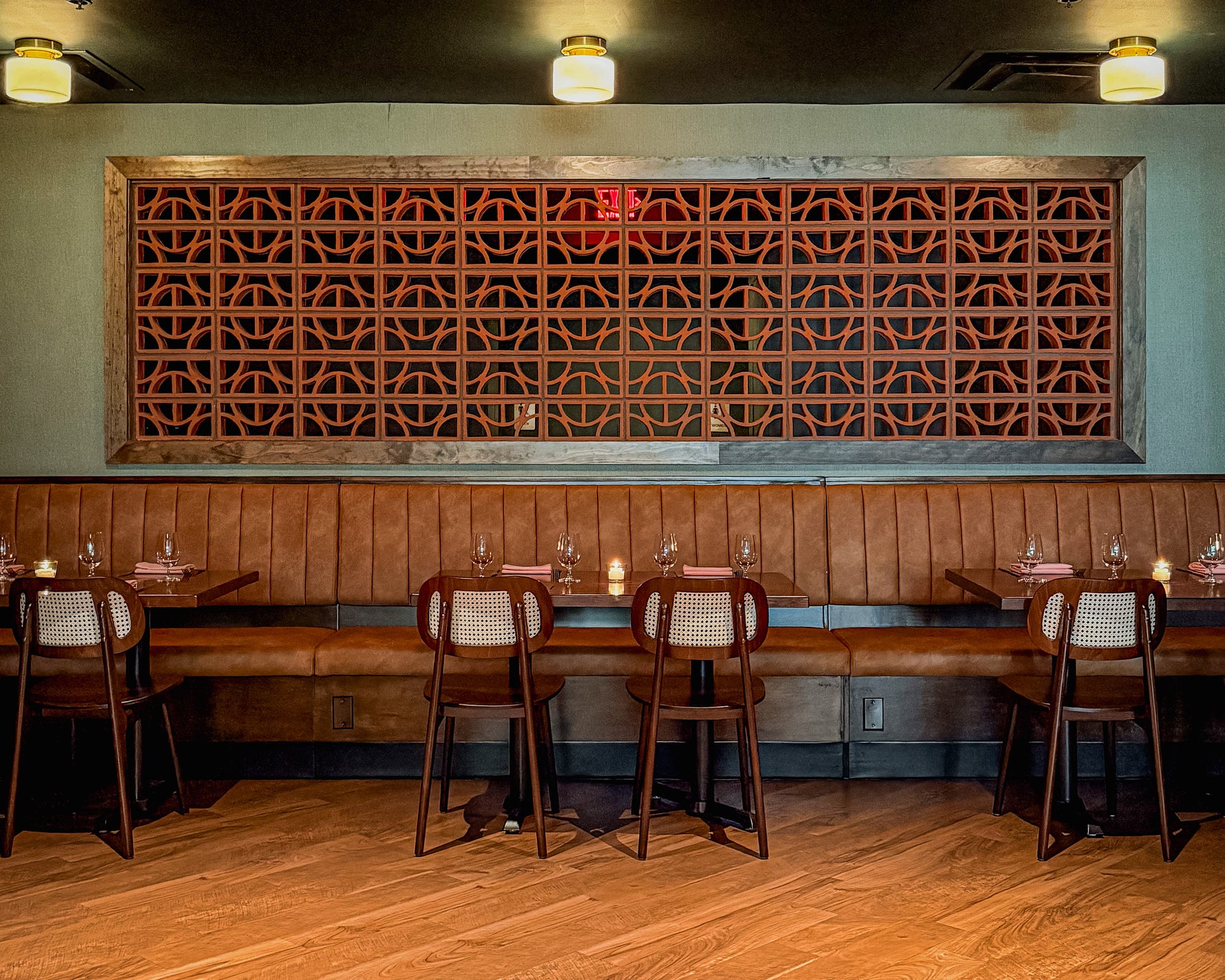 A restaurant interior with a long tan leather booth, dark wood tables, matching chairs with woven backs, and a large decorative geometric wooden wall screen behind the seating.