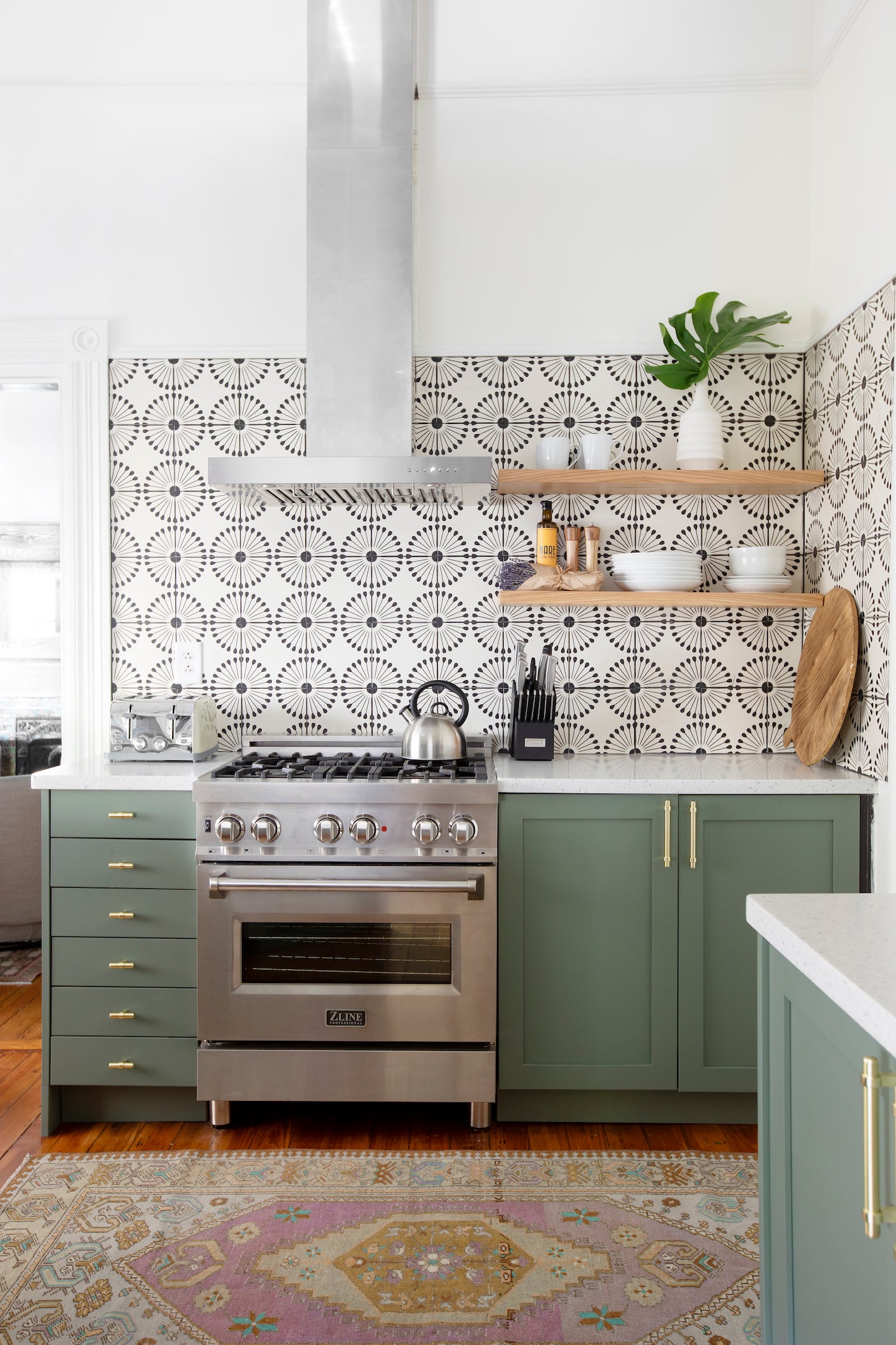 Pitch black 8x8 pattern tile by Atomic Vocho, featuring bold geometric starburst design in black and white, installed as kitchen backsplash with green cabinetry and stainless steel range.